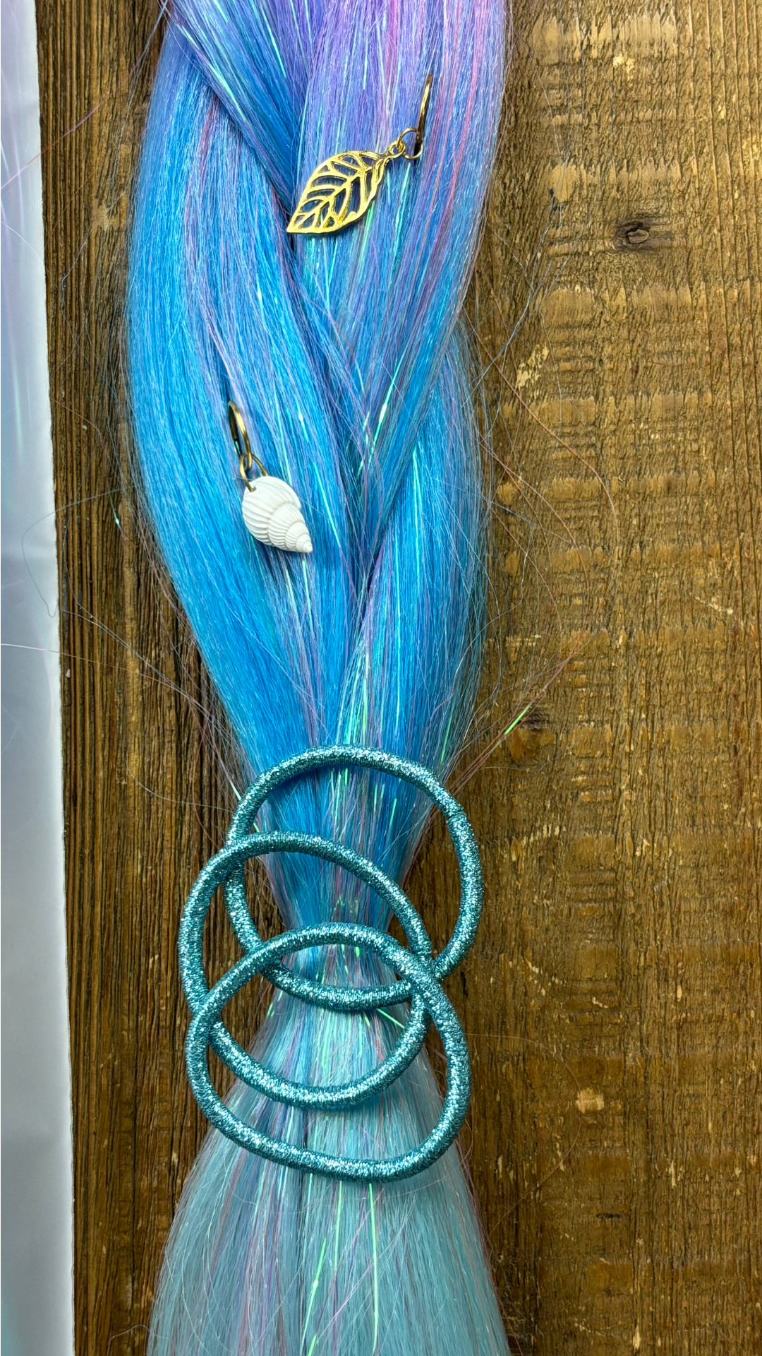 Close up of blue and purple section of rainbow tinsel braid with 3 blue hair ties and golden shell and leaf hair charms on wood background.