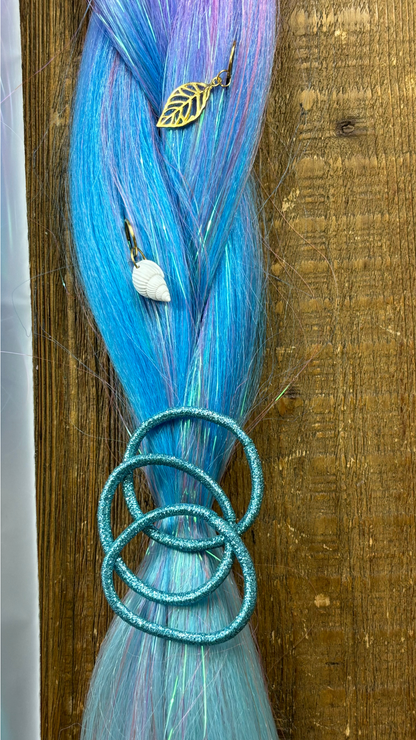 Close up of blue and purple section of rainbow tinsel braid with 3 blue hair ties and golden shell and leaf hair charms on wood background.