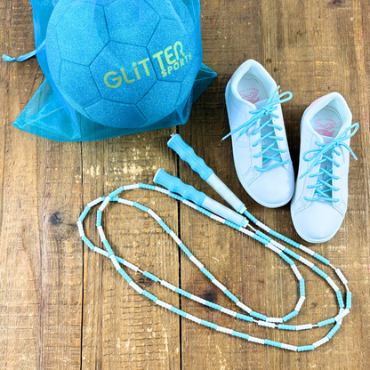 Blue and white soft bead skipping rope places next to white Fila sneakers with holographic blue shoelaces and blue glitter soccer ball with gold 'Glitter Sports' logo inside blue organza gift bag on wood background.