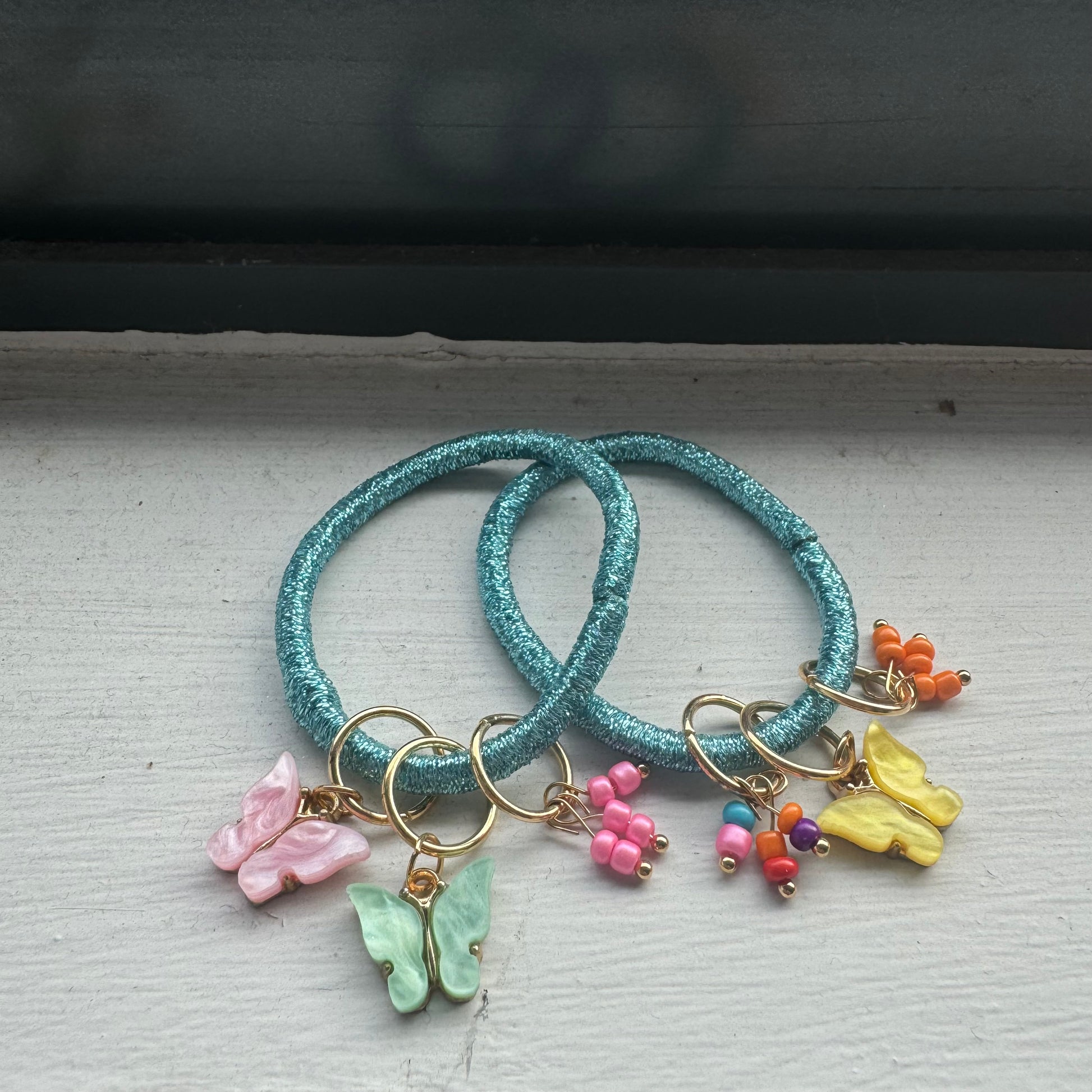 Two metallic blue hair ties with three colourful and gold beaded and butterfly hair charms on each, positioned on white windowsill with black frame.