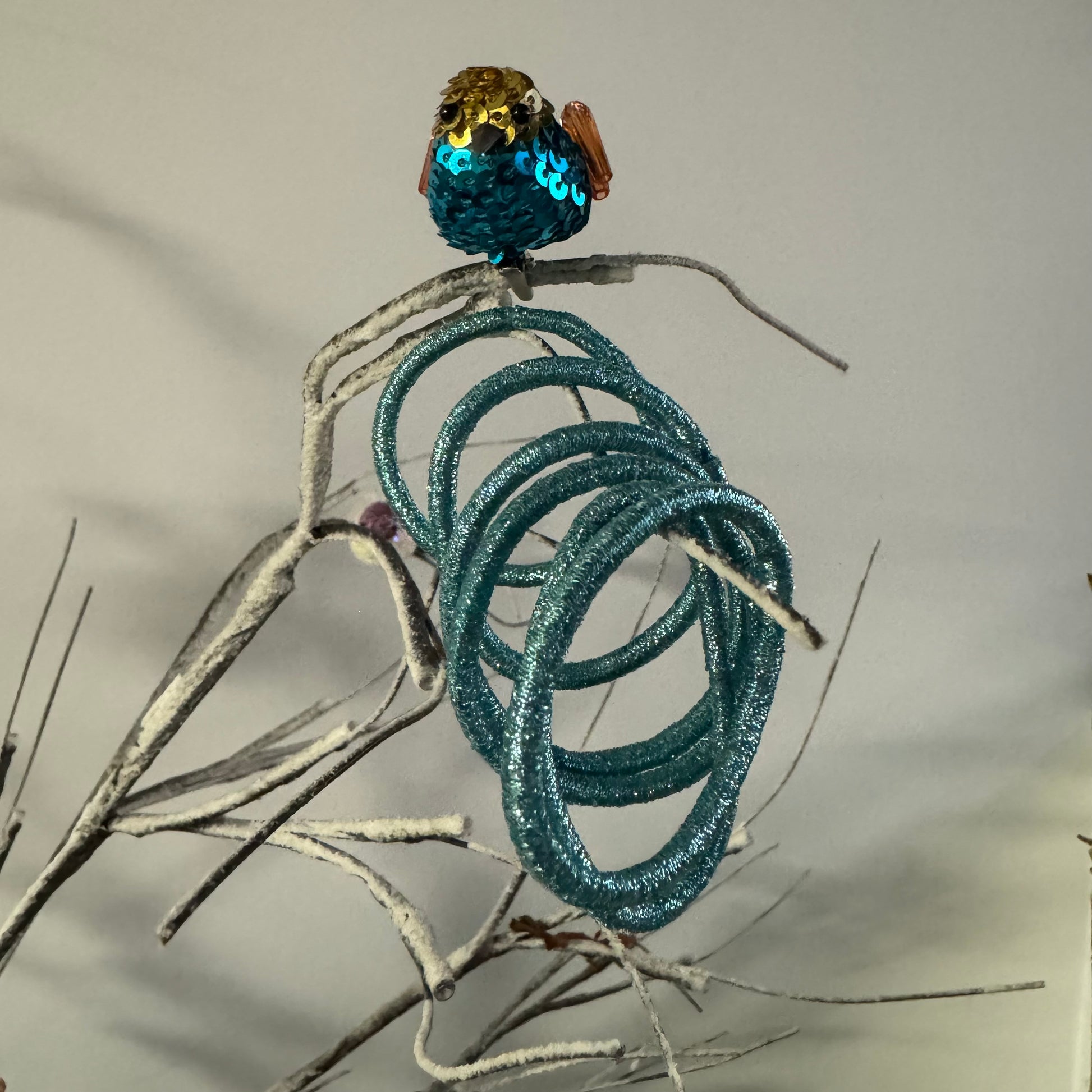 6 metallic blue hair elastics positioned on grey branch next to small sequin bird decoration on white background.