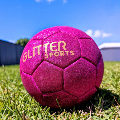 Bright pink glitter soccer ball with gold 'Glitter Sports' logo sitting on grass in backyard, sparkling in the sun.