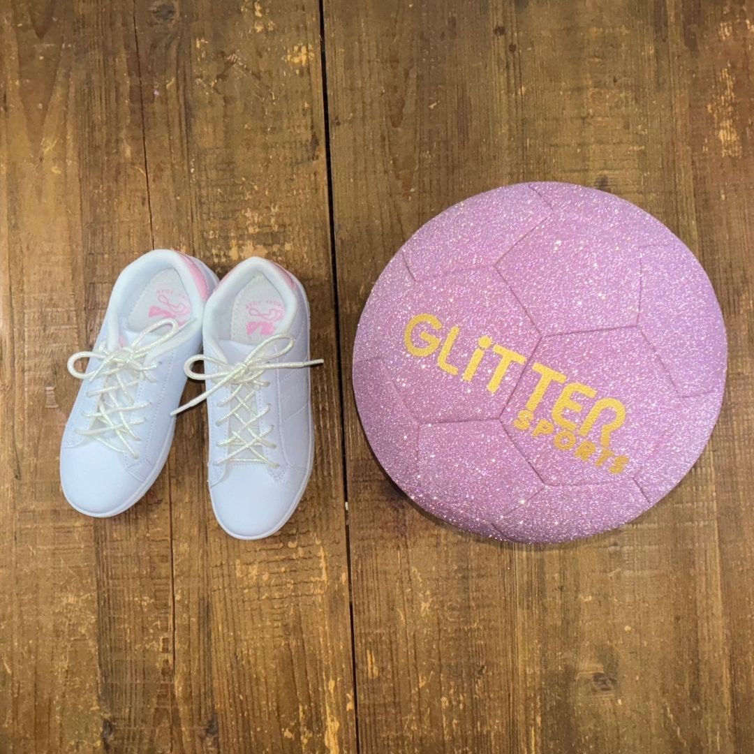 Girls white and pink Fila sneakers with white pearlescent shoelaces next to sparkly peony pink soccer ball with gold 'Glitter Sports' logo on wood background.