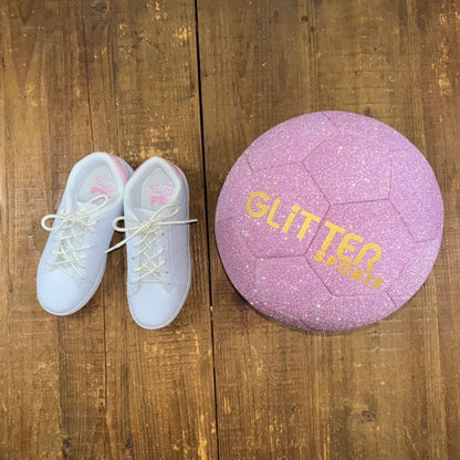 Girls white and pink Fila sneakers with white pearlescent shoelaces next to sparkly peony pink soccer ball with gold 'Glitter Sports' logo on wood background.