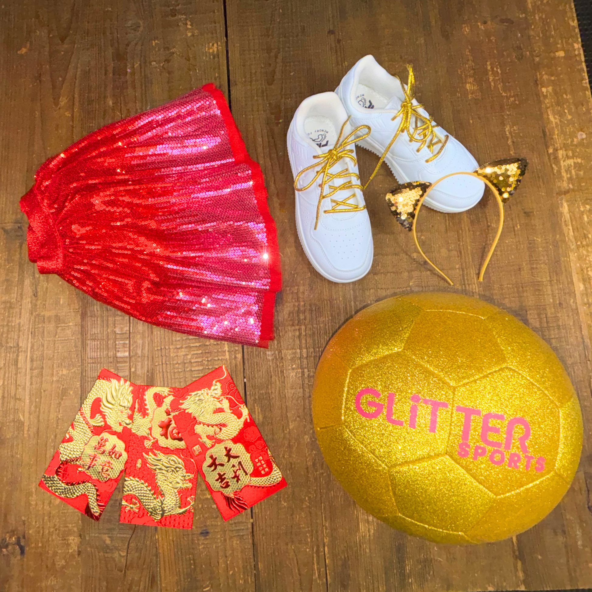 Overhead photo of size 3 gold glitter kids soccer ball, CNY red dragon envelopes, white sneakers with gold metallic shoelaces, sequin headband, red and pink girls tutu laid on wooden table.