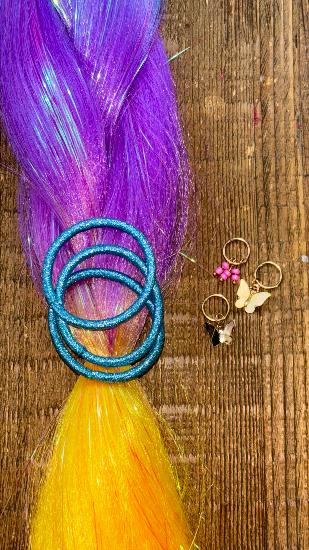 Magenta purple and golden yellow section of rainbow tinsel braids with aqua metallic hair ties and golden butterfly hair charms on wooden background.