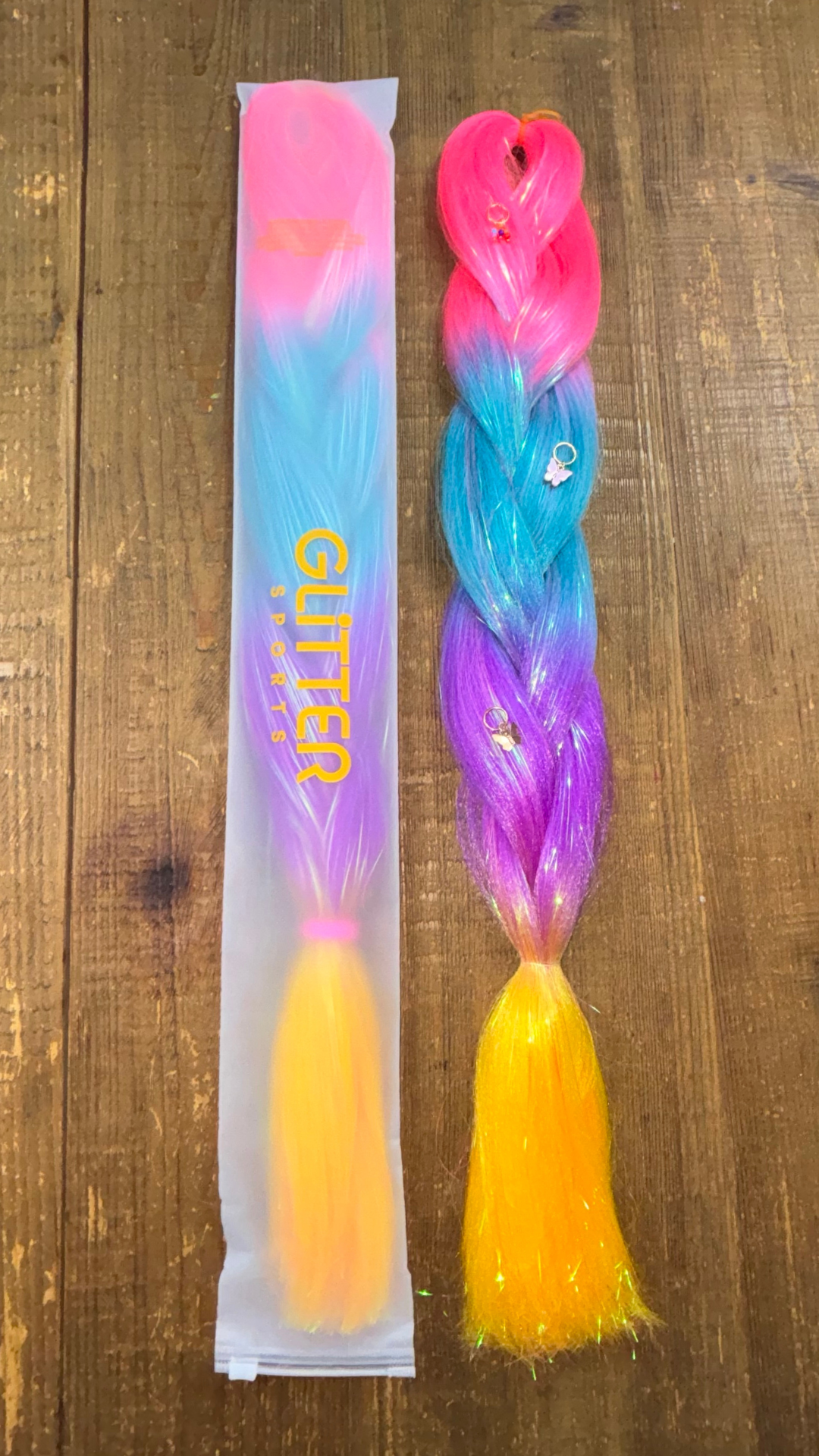 Rainbow tinsel synthetic hair extension braids with golden butterfly hair charms on wooden background. Braid colours ombre through fuchsia, aqua, magenta purple and golden yellow.