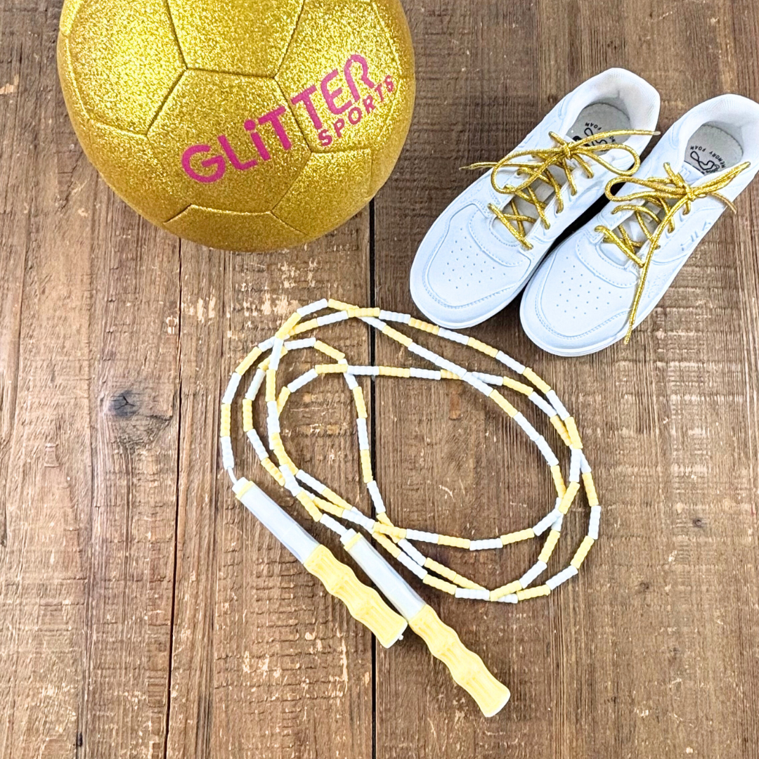 Lemon yellow and white soft bead skipping rope places next to white Reebok joggers with metallic gold round shoelaces and 24 carat gold glitter soccer ball with pink 'Glitter Sports' logo on wood background.