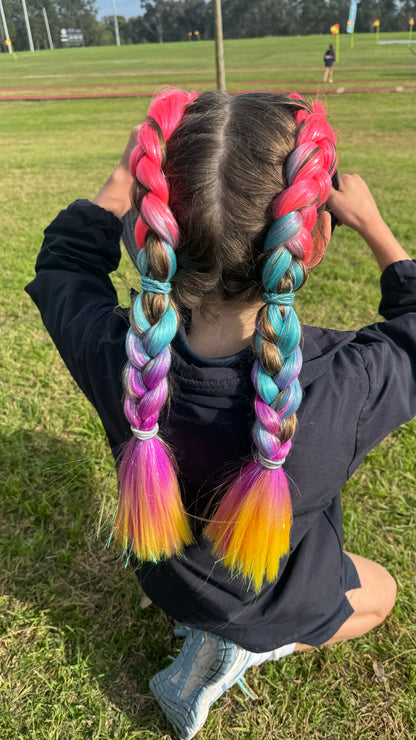 Young girl at park facing away to show two rainbow tinsel extension braids in her hair with ombre of fuchsia, blue, magenta and golden yellow.