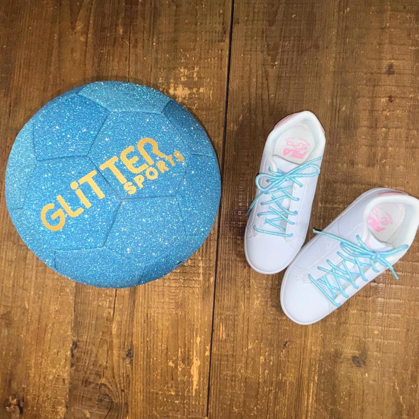 Blue sparkly glitter soccer ball with gold 'Glitter Sports' logo next to kids white and pink Fila sneakers with blue shoelaces on wood background.