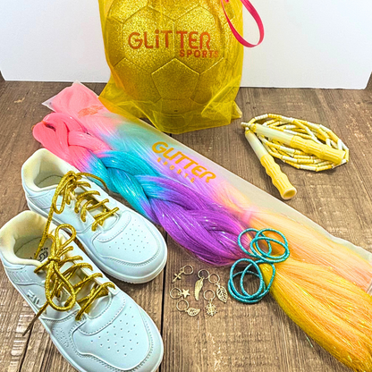 White Fila sneakers with metallic gold round shoelaces next to rainbow synthetic hair extension braids with pink, blue purple and golden yellow and tinsel running through with blue metallic hair elastics and assorted gold hair charms. Lemon yellow and white soft bead skipping rope placed next to gold glitter soccer ball with pink 'Glitter Sports' logo inside gold organa gift bag on wood table with white background.