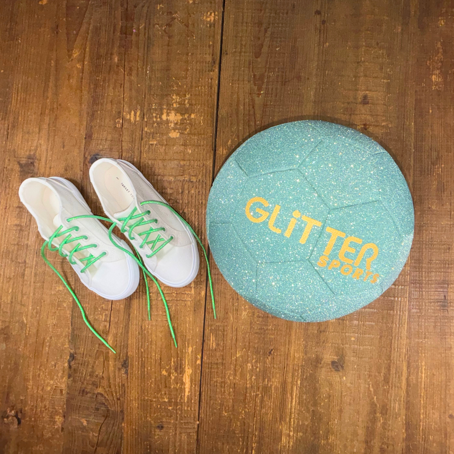 Two white kids canvas sneakers from Target with bright green metallic shoelaces next to a mint green glitter soccer ball with gold 'Glitter Sports' logo on wood background.