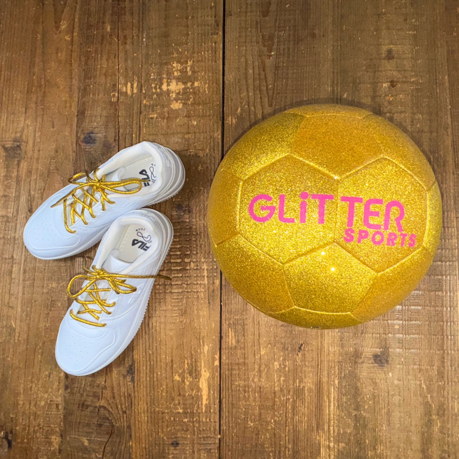 White Fila kids sneakers with metallic gold shoelaces next to gold glitter soccer ball with pink 'Glitter Sports' logo on wood background.