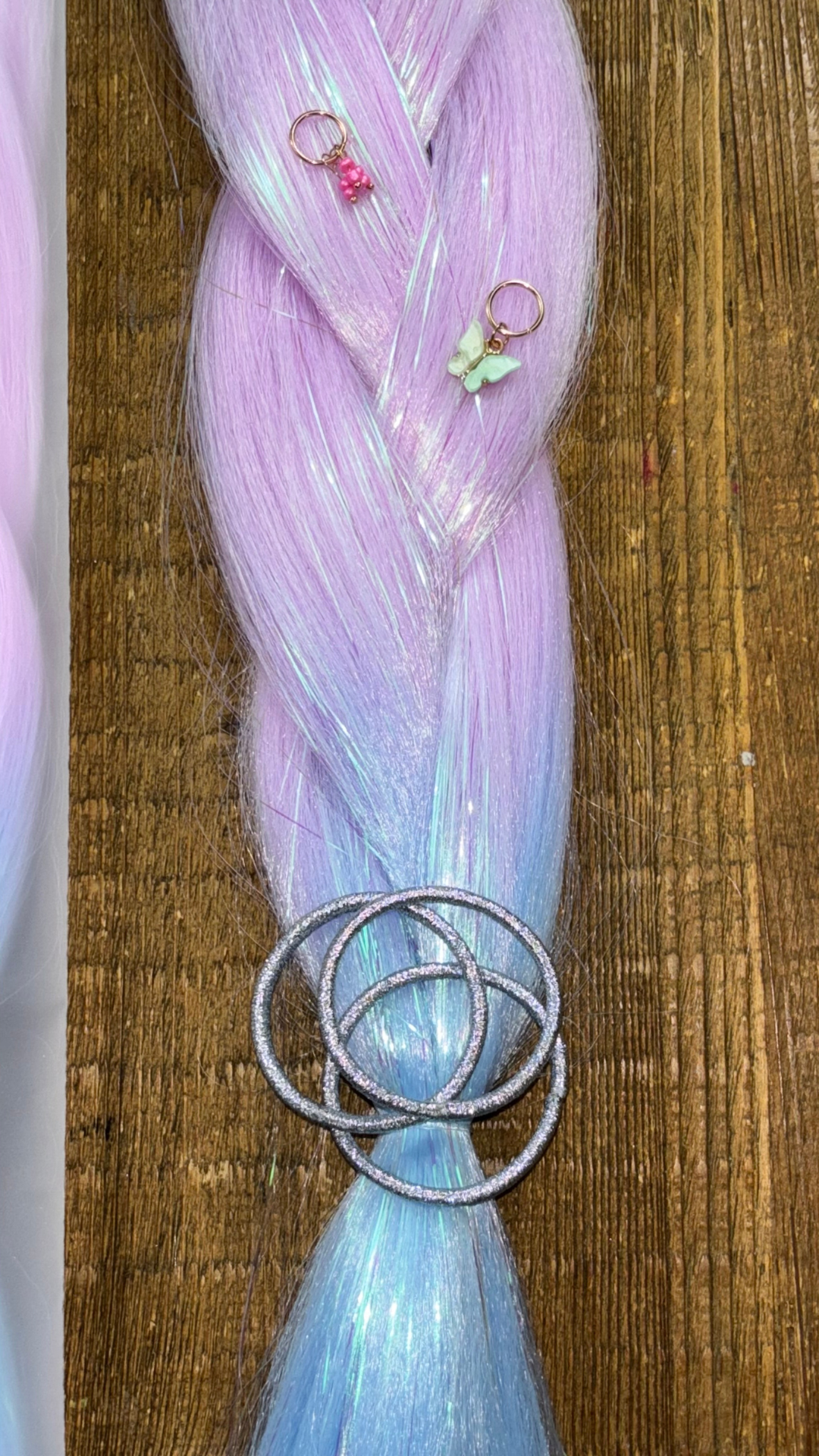 Ombre tinsel hair extension braid in lavender and blue with silver metallic hair elastics and pink and green butterfly and bead hair charms on wood background.