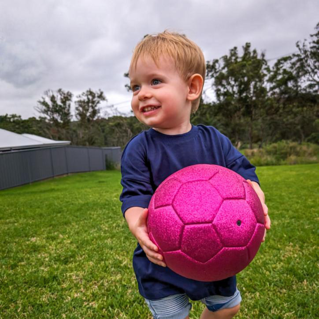 Cute little boy in navy shirt and striped shorts smiles and looks to his right while holding pink glitter soccer ball and standing in back yard.