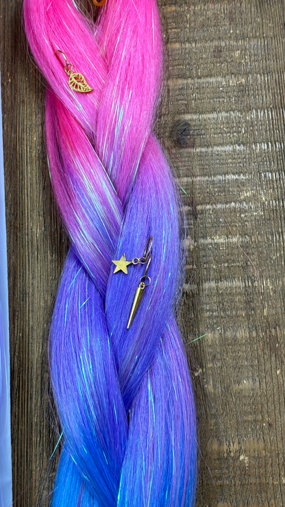 Close up of pink and purple rainbow tinsel hair extension braid with gold hair charms in star and leaf on wood background.