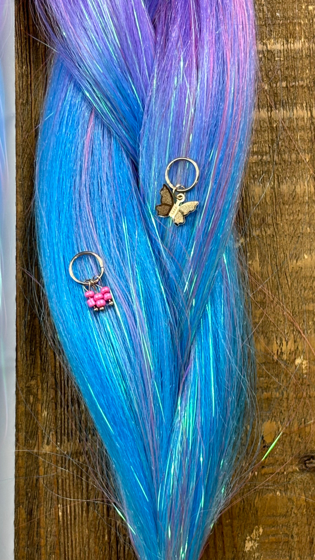 Close up of purple and blue ombre tinsel hair extension braid with pink and gold butterfly hair charms on wood background.