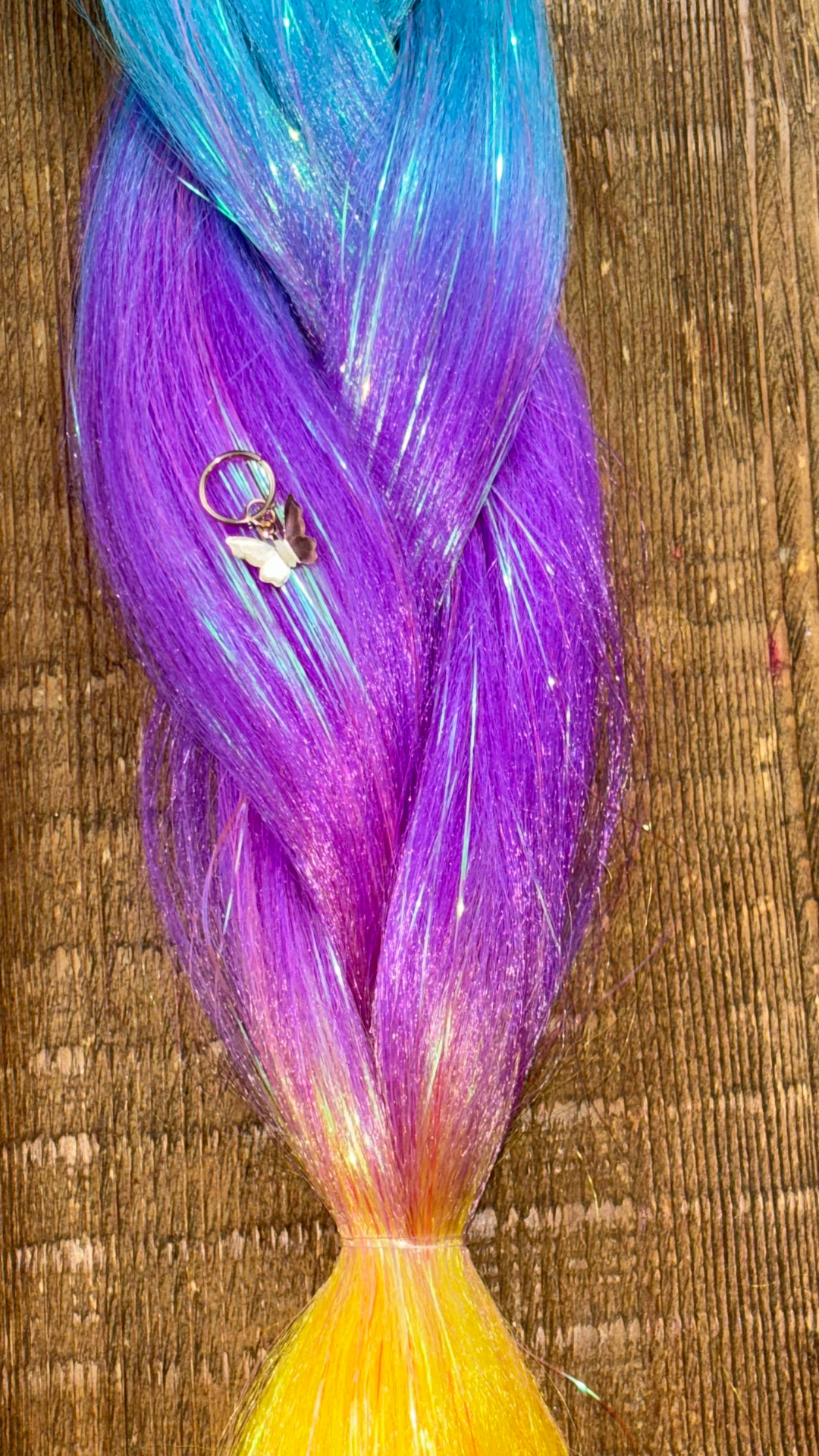 Rainbow ombre tinsel braids close up of blue, magenta purple and golden yellow section with gold butterfly hair charm on wooden background.