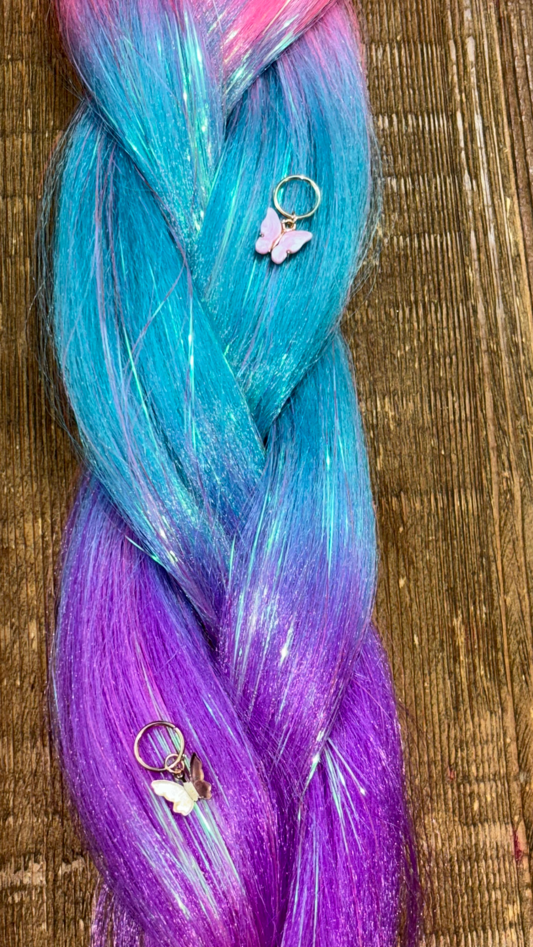 Close up of blue, pink, purple sections of rainbow tinsel braids with pink and gold butterfly hair charms on wooden background.