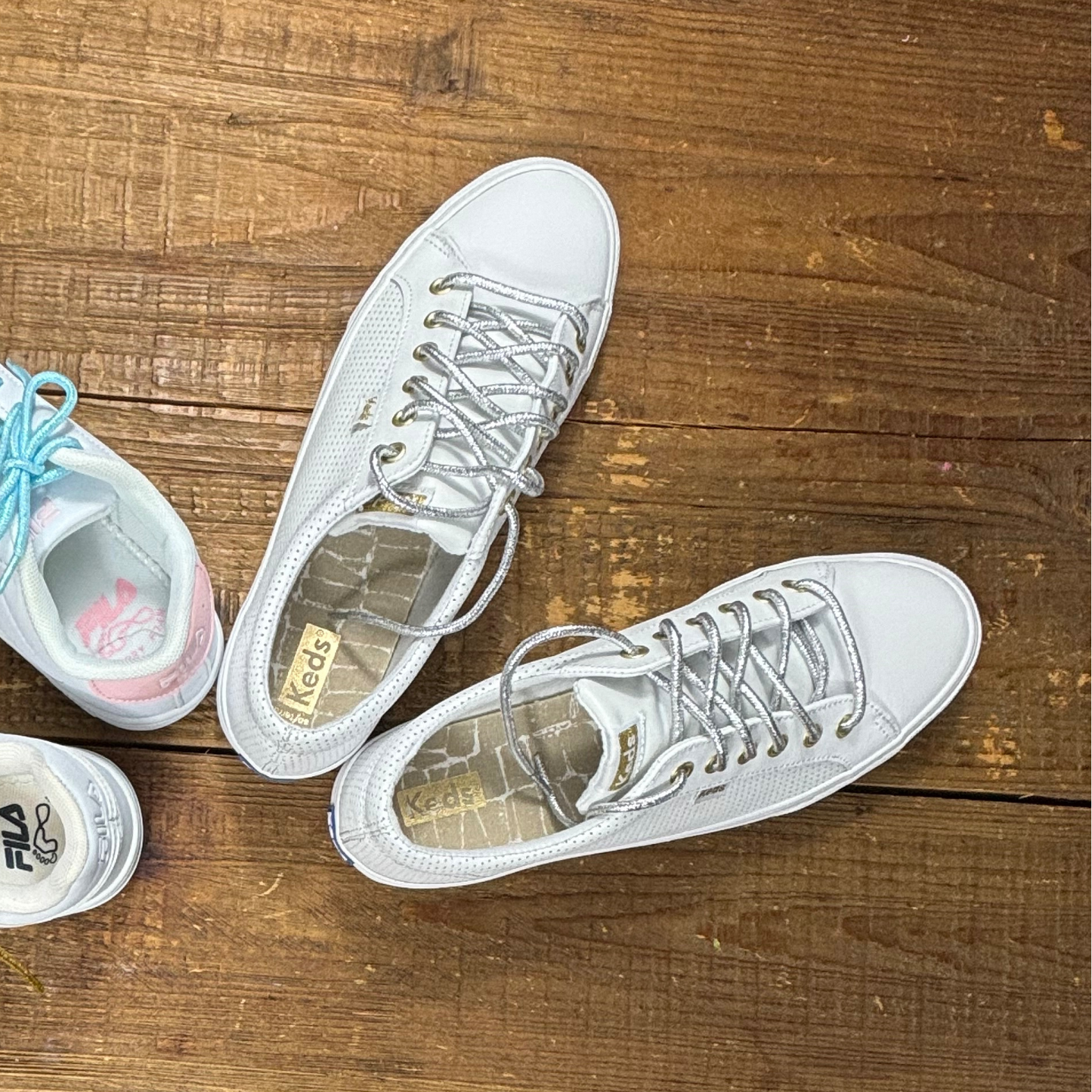 White Keds leather sneakers with metallic silver lurex shoelaces next to white and pink Fila sneaker with blue shoelaces on wood background.