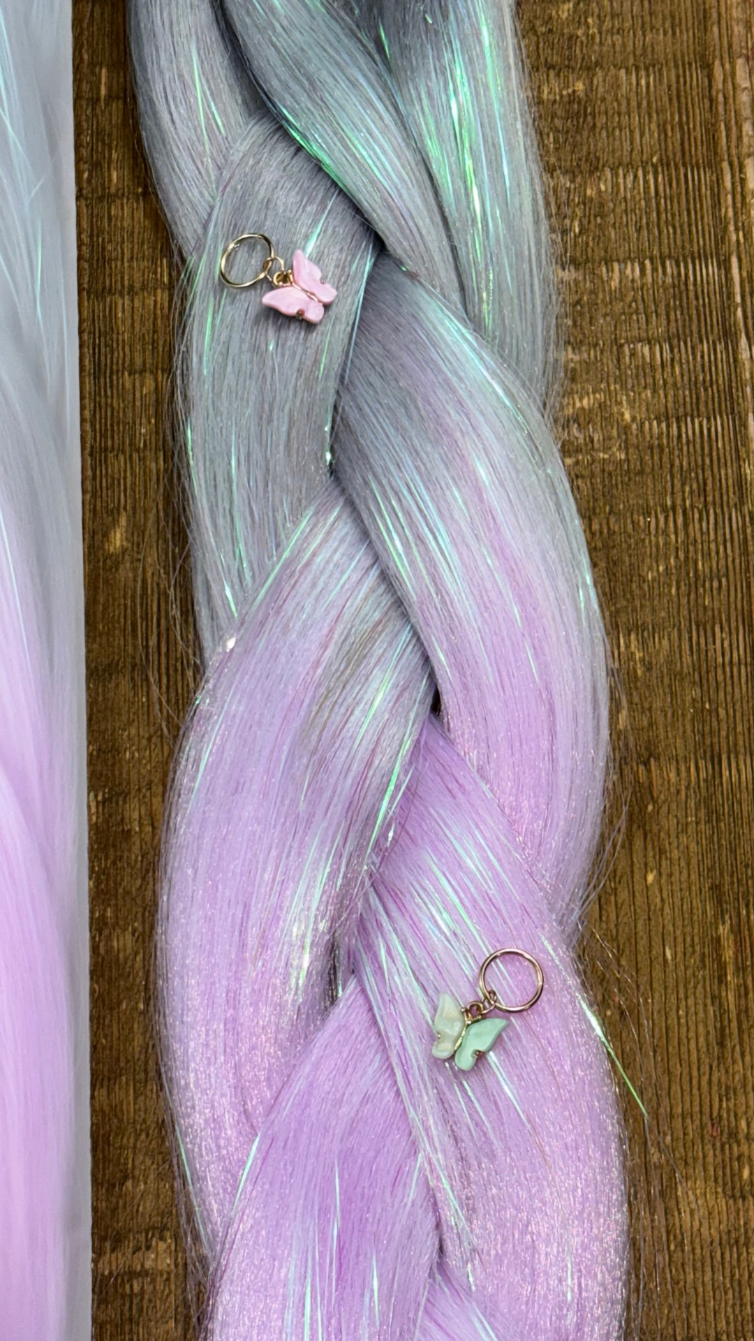 Close up of silver and purple synthetic hair extension braids with pink and green hair charm butterflies on wood background.