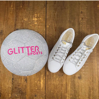 Metallic silver round shoelaces in white leather Keds sneakers next to diamond glitter soccer ball with pink 'Glitter Sports' logo on wood background.