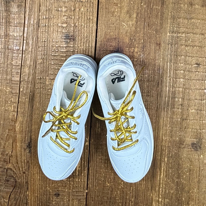 White kids Fila sneakers with metallic gold round shoelaces on wood background.