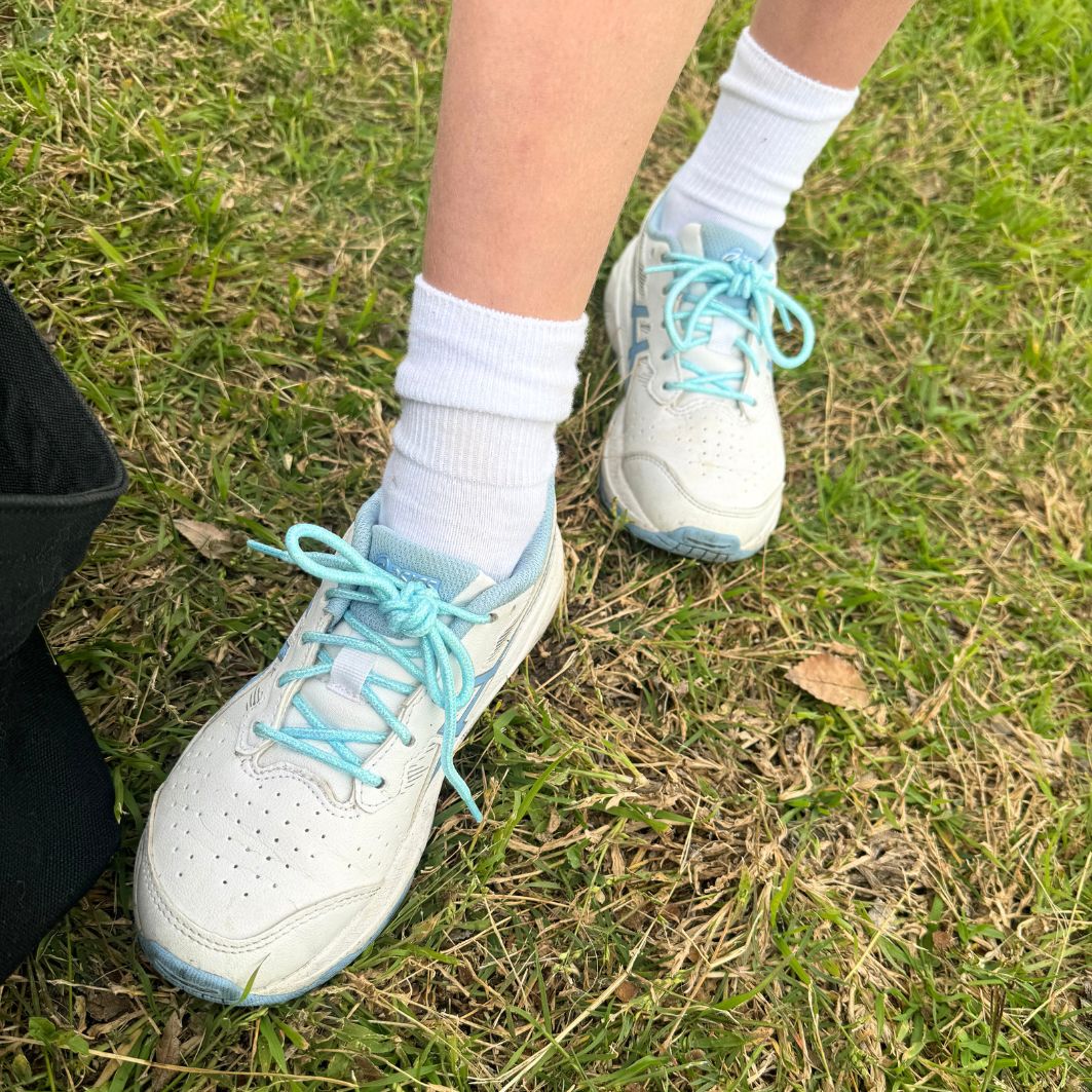 Kids lower legs wearing white socks and white and blue sneakers with blue shoelaces standing on grass.