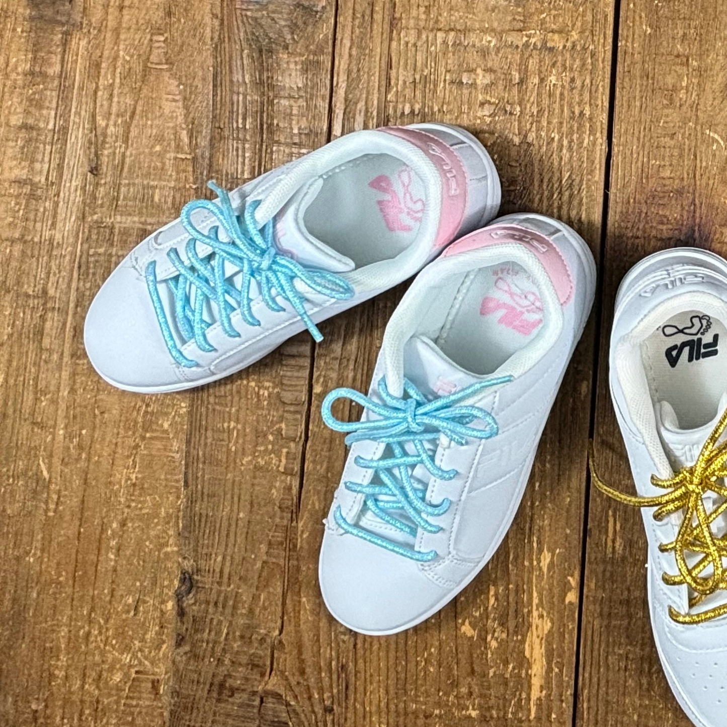 White and pink fila sneakers with blue shoelaces next to white file sneaker with gold shoelaces on wood background.