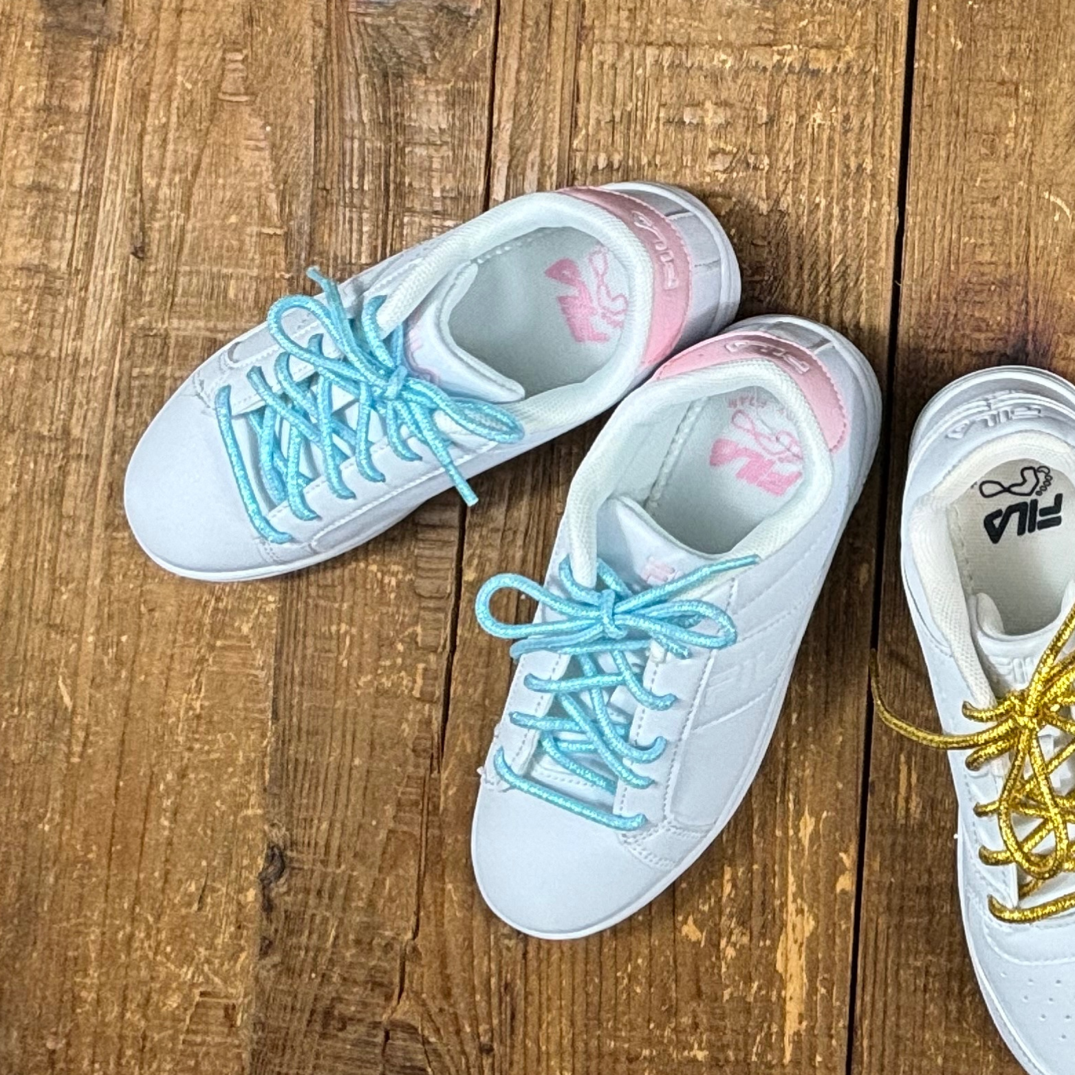 White and pink fila sneakers with blue shoelaces next to white file sneaker with gold shoelaces on wood background.