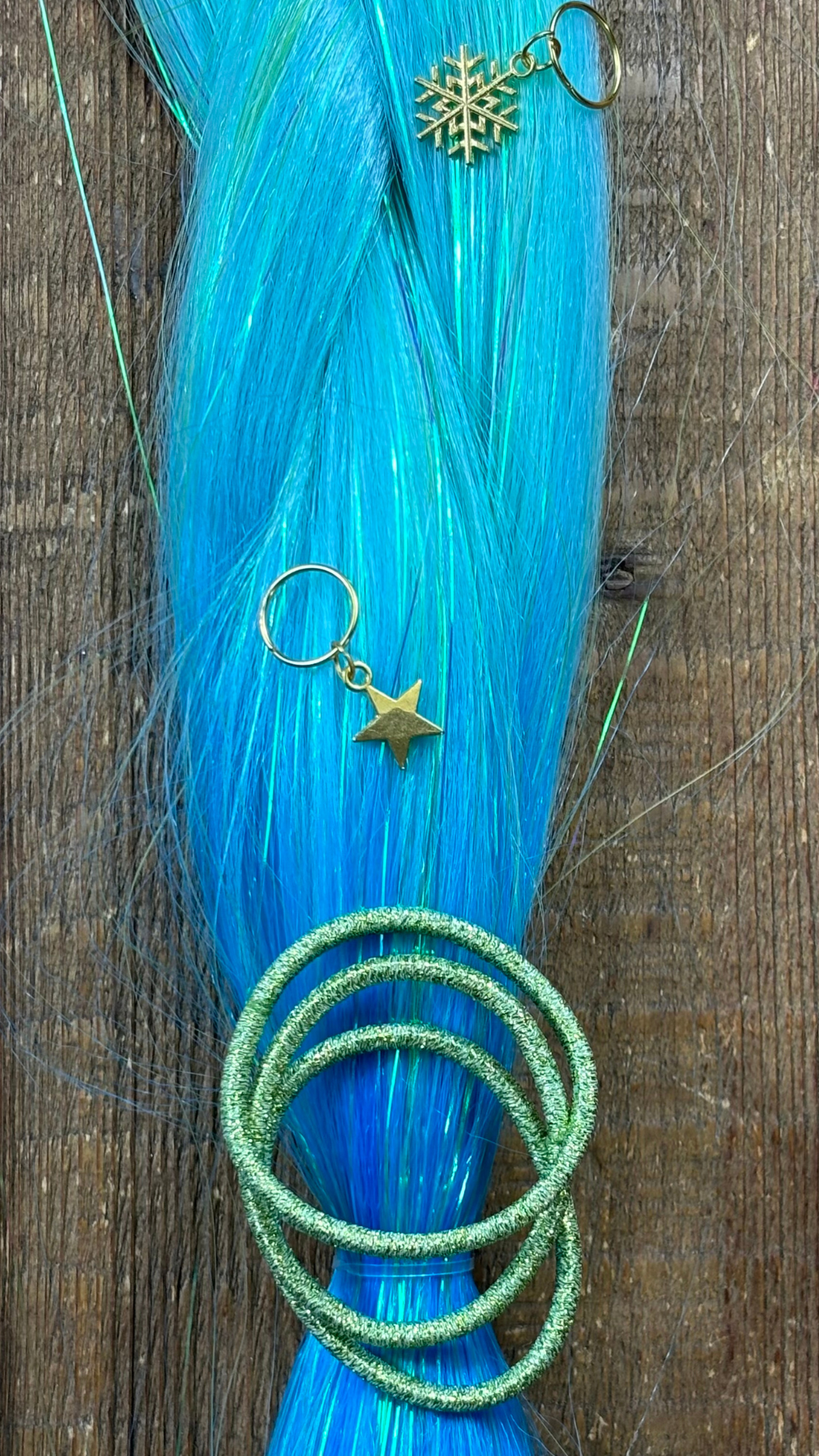 Closeup of aqua blue tinsel extension braids with metallic peridot hair ties and gold snowflake and star hair charms on wood background.