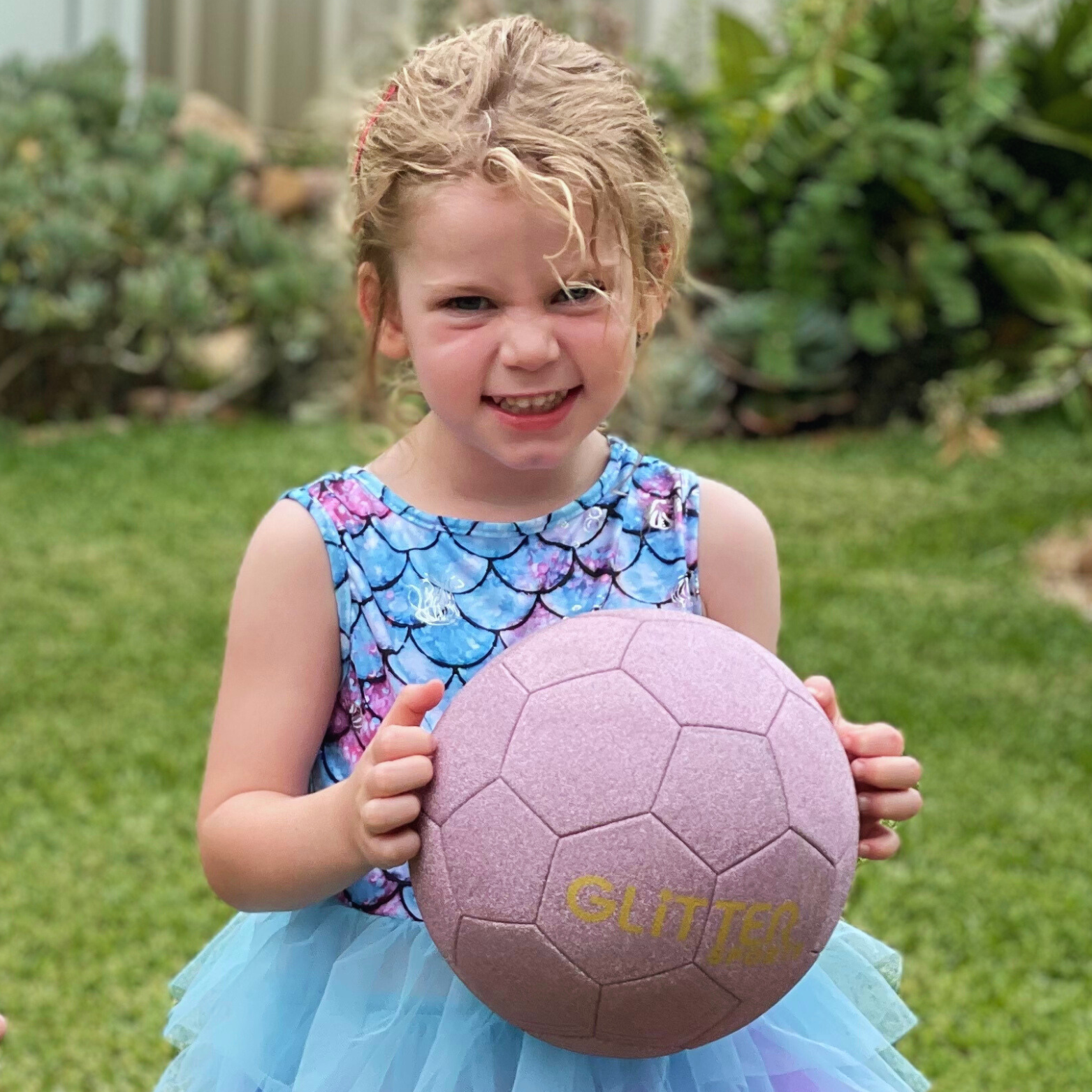 Young girl with mischievous expression wearing blue and purple dress with mermaid pattern on top and blue tule skirt holds pink glitter soccer ball with garden backyard background.