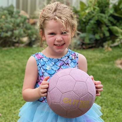 Young girl with mischievous expression wearing blue and purple dress with mermaid pattern on top and blue tule skirt holds pink glitter soccer ball with garden backyard background.