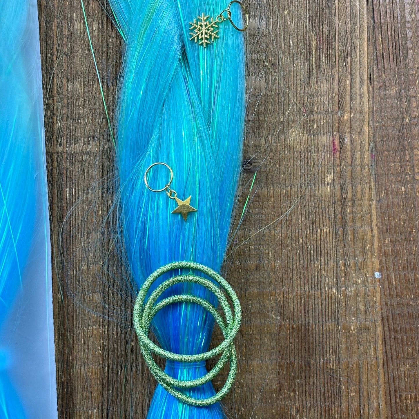 Closeup of blue hair extension with tinsel running through, green metallic hair ties and gold star and snowflake hair charms on wood background.