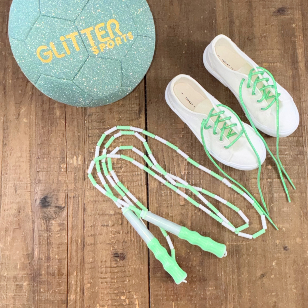 Mint green and white soft bead skipping rope next to white Target canvas sneakers with green metallic shoelaces and mint glitter kids soccer ball with gold'Glitter Sports' logo on wood background.