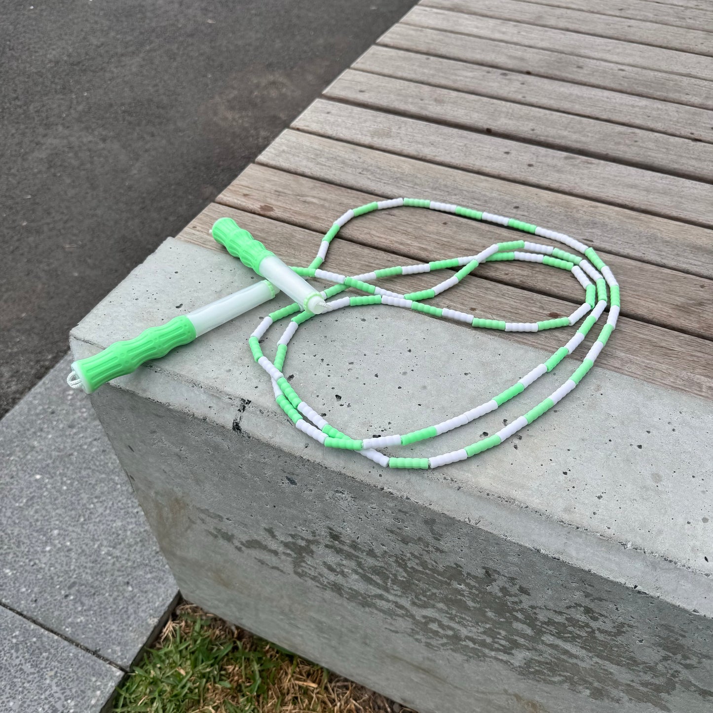 Mint green and white soft bead skipping rope places on concrete and wood bench next to grass and pathway