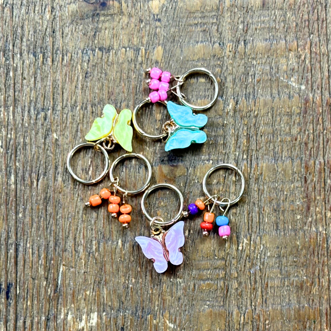 Six hair charms on gold hoops including colourful butterflies and beads in pink, yellow and green on wood background.