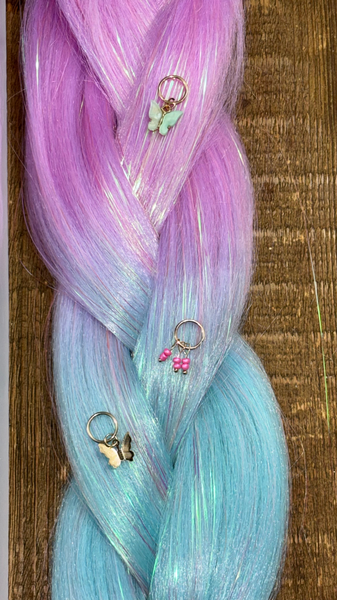 Close up of tinsel synthetic braid extension with pink and blue ombre and butterfly hair charms and beads on wood background.