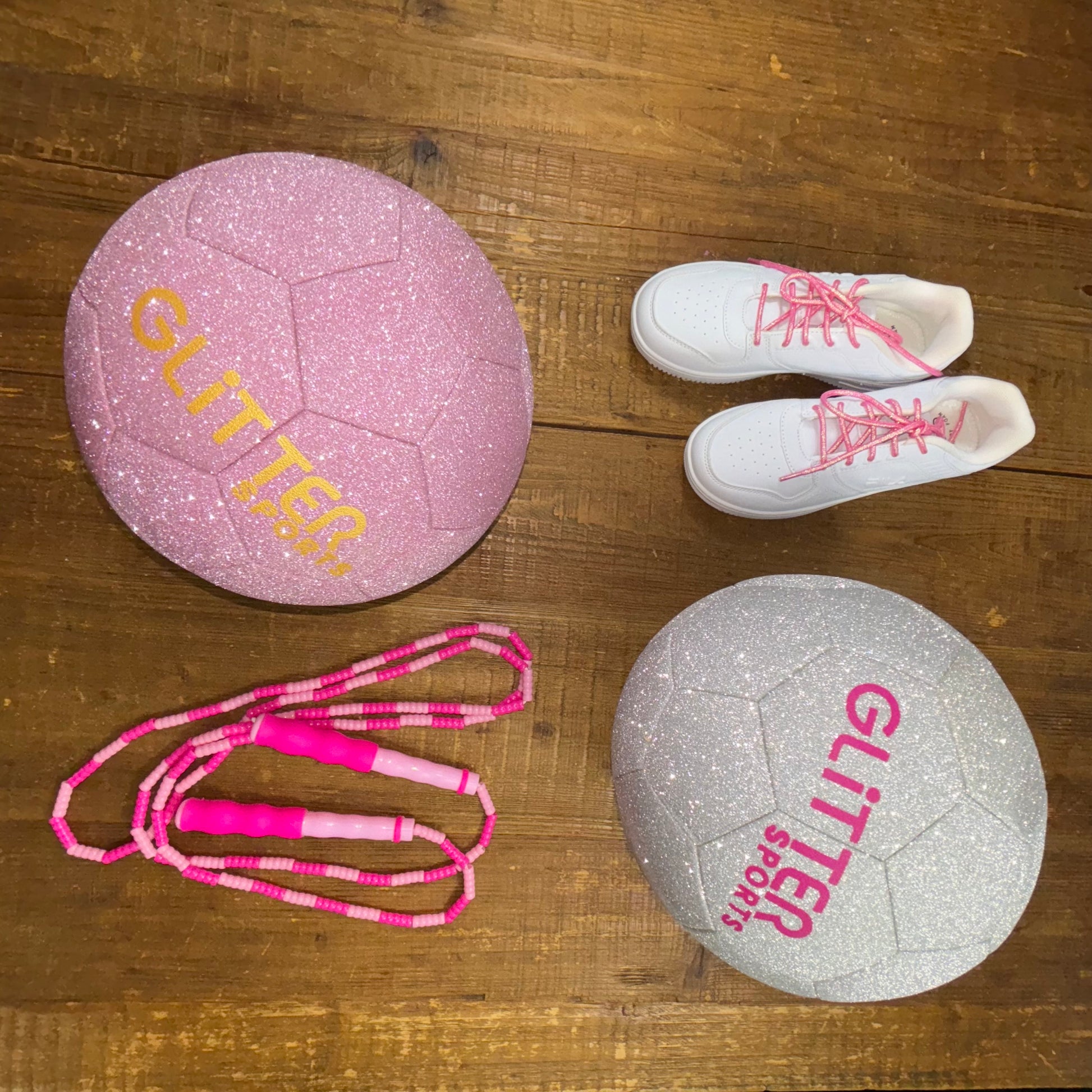 Two glitter soccer balls in pink and silver diamond with 'Glitter Sports' logos next to pink two-toned soft bead skipping rope and white reebok sneakers with pink round metallic shoelaces on wood background.