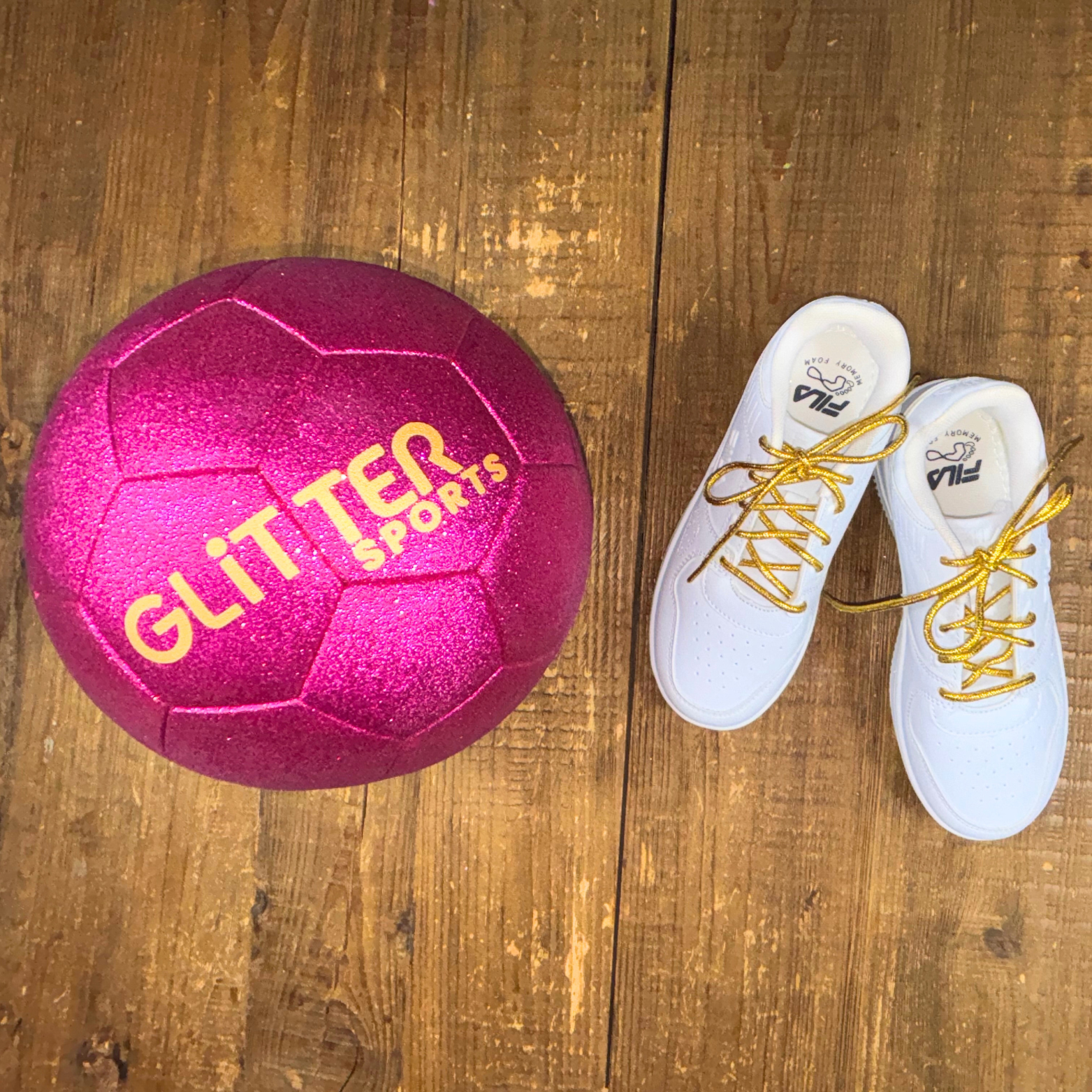 Bright pink glitter soccer ball with gold 'Glitter Sports' logo next to girls white Fila sneakers with metallic gold shoelaces on wood background.