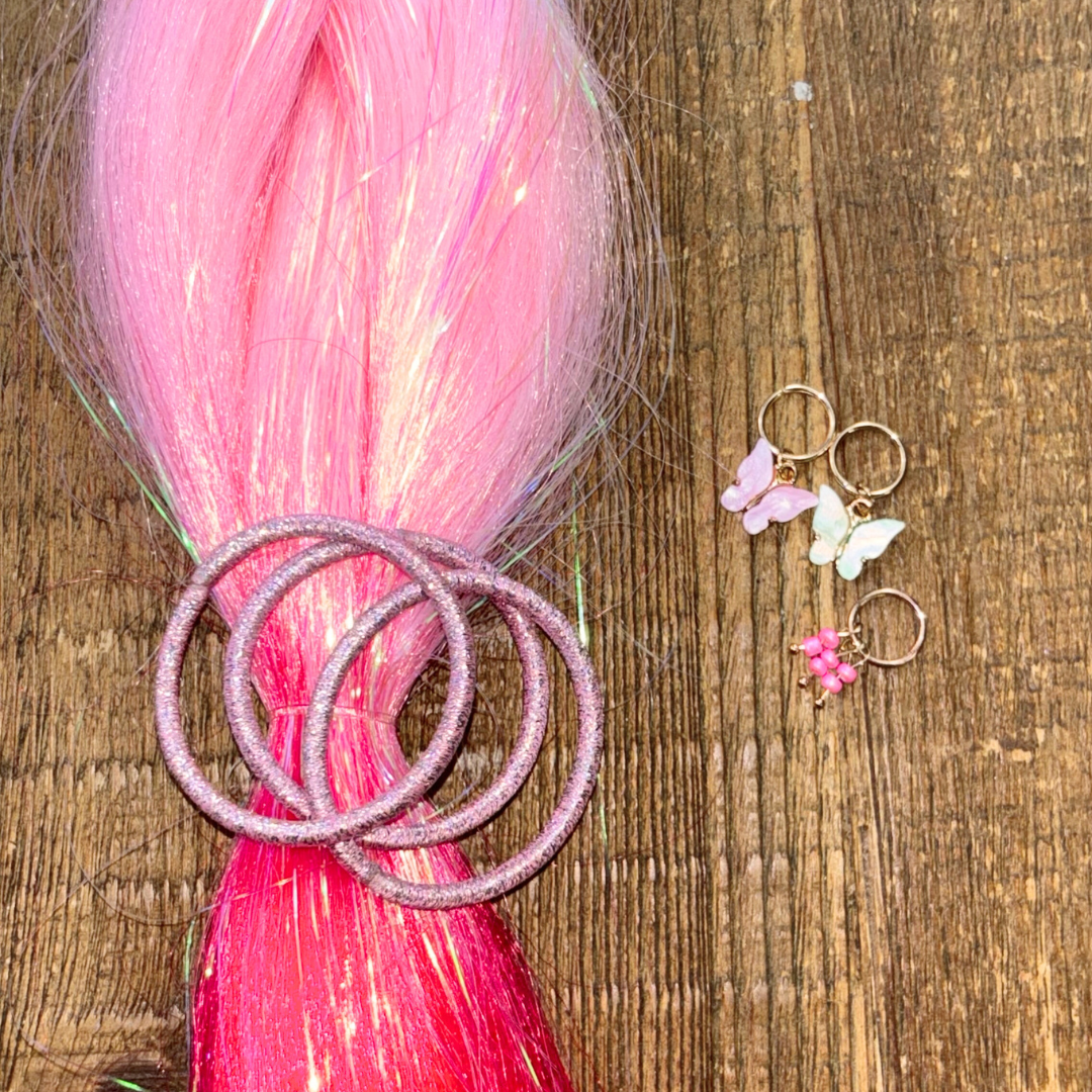 Closeup of ombre pink tinsel hair extension with 3 pink metallic hair elastics and 3 pink butterfly and bead gold hair charms on wood background.