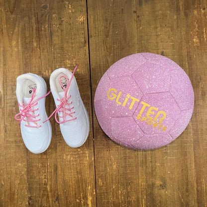 White girls fila sport sneakers with pink metallic shoelaces next to pink sparkly glitter soccer ball with gold 'Glitter Sports' logo on wood background.