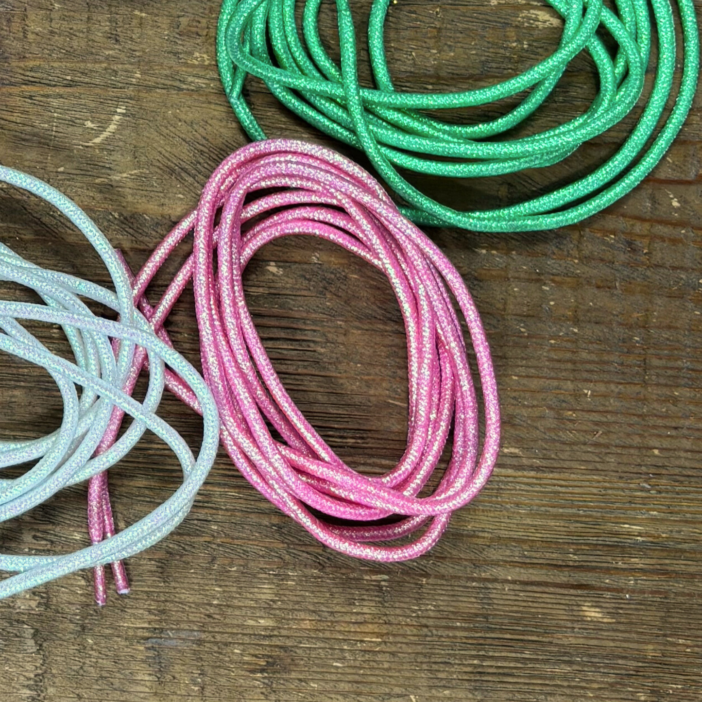 Pink metallic shoelaces placed in a circle on a wood background next to green metallic and white pearlescent shoelaces.