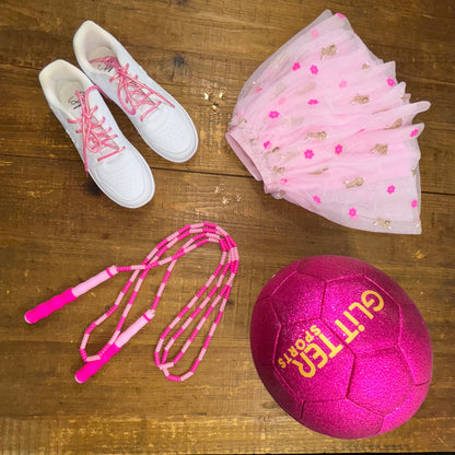 Two-toned pink soft bead skipping rope next to bright pink glitter soccer ball with gold 'Glitter Sports' logo, pink Easter tutu with sequin flowers and rabbits and white reebok sneakers with pink metallic shoelaces on wood background.