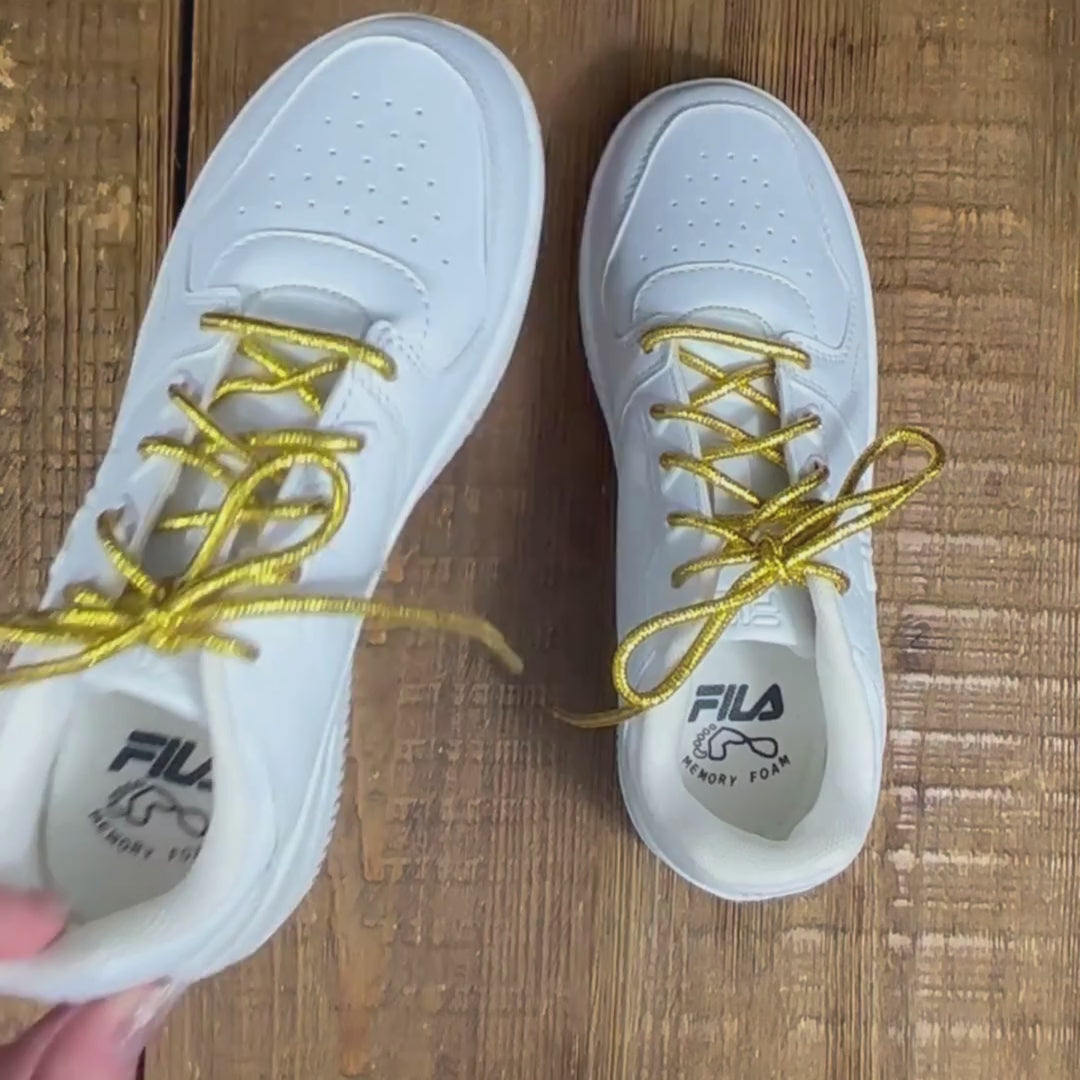 Close up of kids white Fila joggers with metallic 'Stay Gold' shoelaces on wood background.