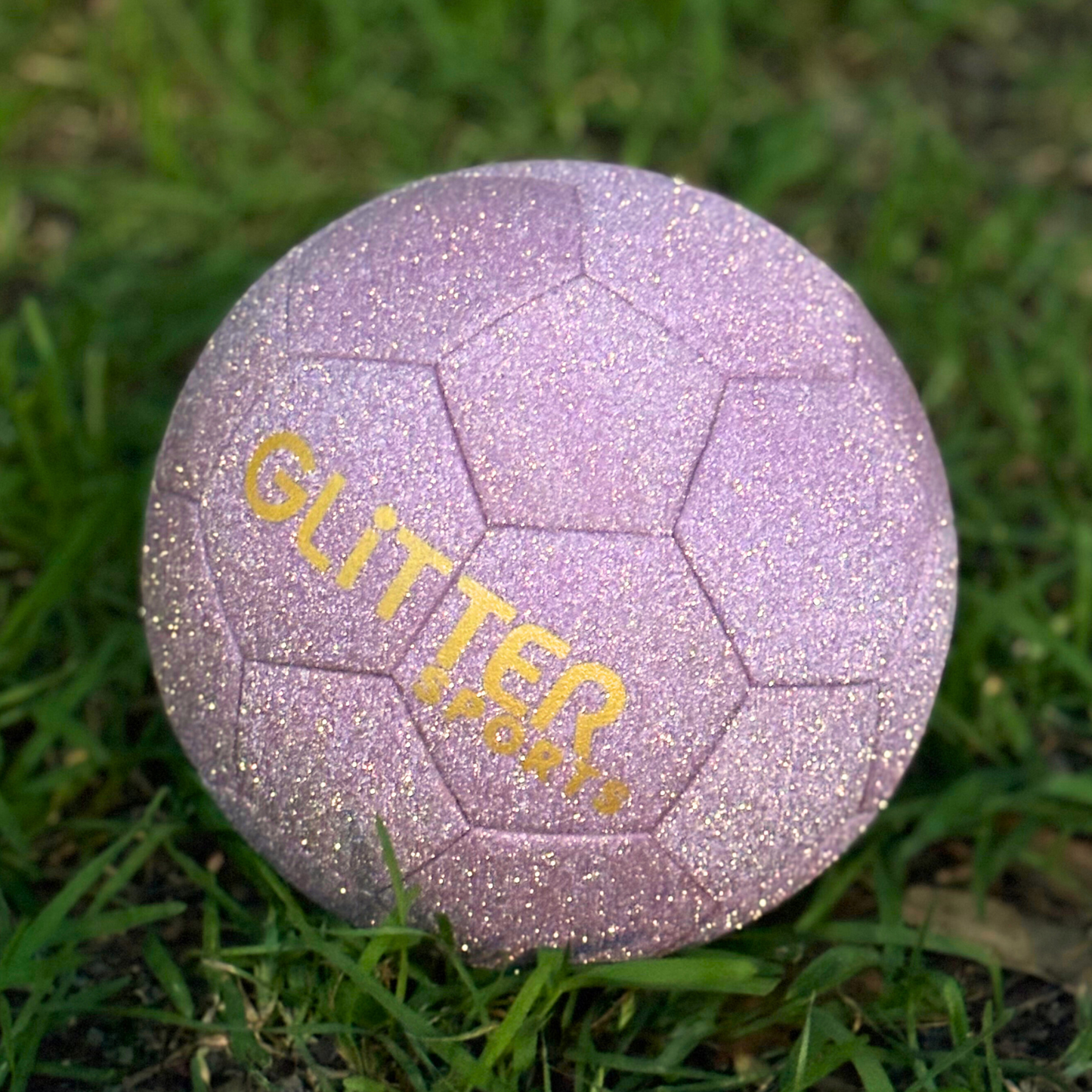 Super sparkly pink glitter soccer ball with gold 'Glitter Sports' logo sits on bright green grassy.