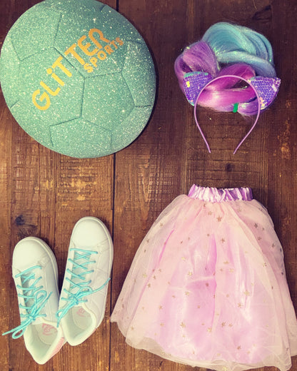 Size 3 Spearmint green glitter soccer ball with gold 'Glitter Sports' logo next to white Fila sneakers with metallic blue shoelaces, purple girls tutu with gold stars, tinsel hair extension braids and purple sequin kitten ears hairband on wood background.