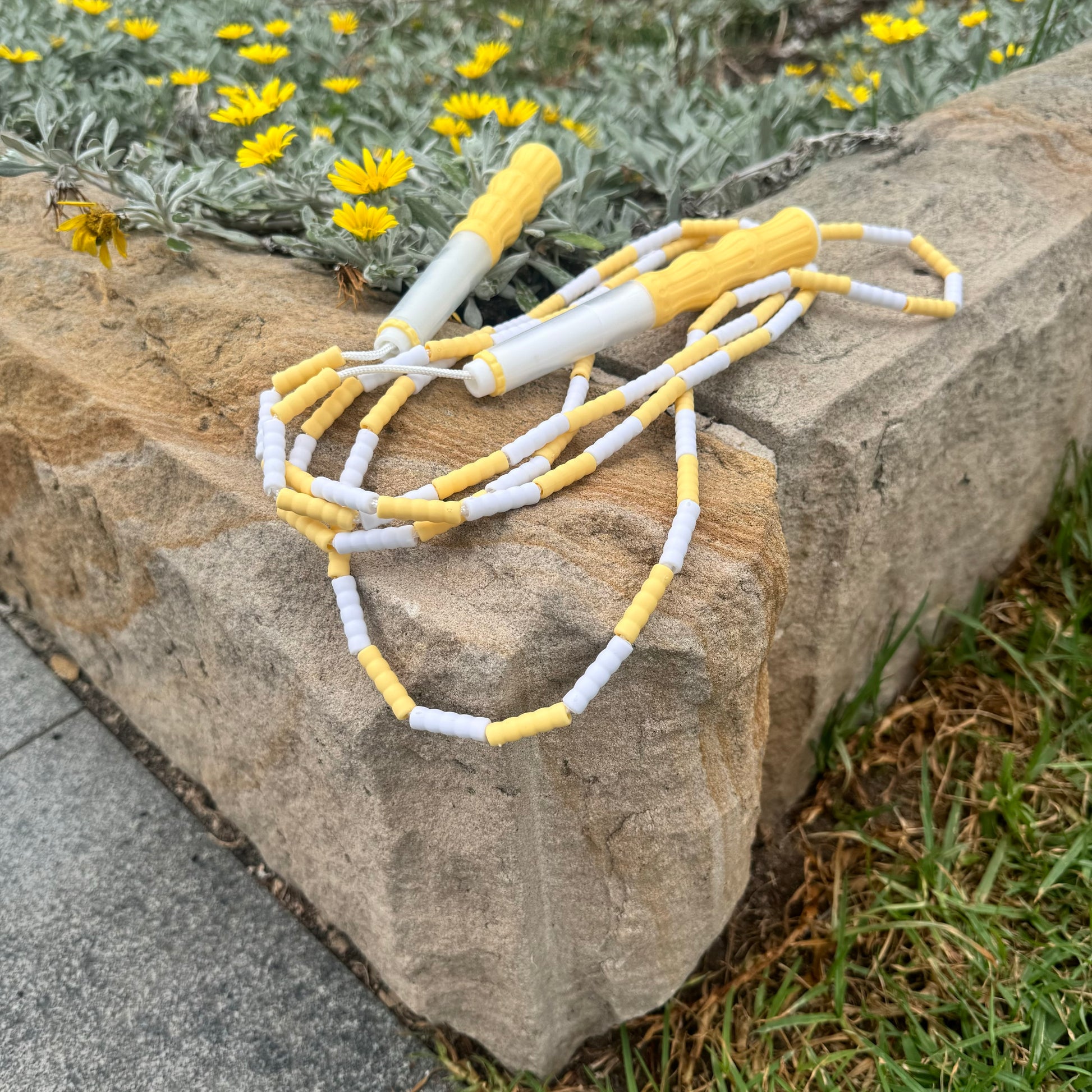 Lemon yellow and white soft beed skipping rope placed on sandstone garden border with yellow daisies in background and grass and pathway in front.